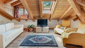 A cozy attic room with wooden beams, a large window, a flat-screen TV, a white sofa, a yellow armchair, and a patterned rug.