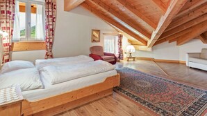 A bedroom with a sloped ceiling, wooden beams, a bed, a chair, a lamp, and a view through a window.