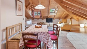 A cozy attic room with a wooden ceiling, a dining table with chairs, a sofa, and a TV.