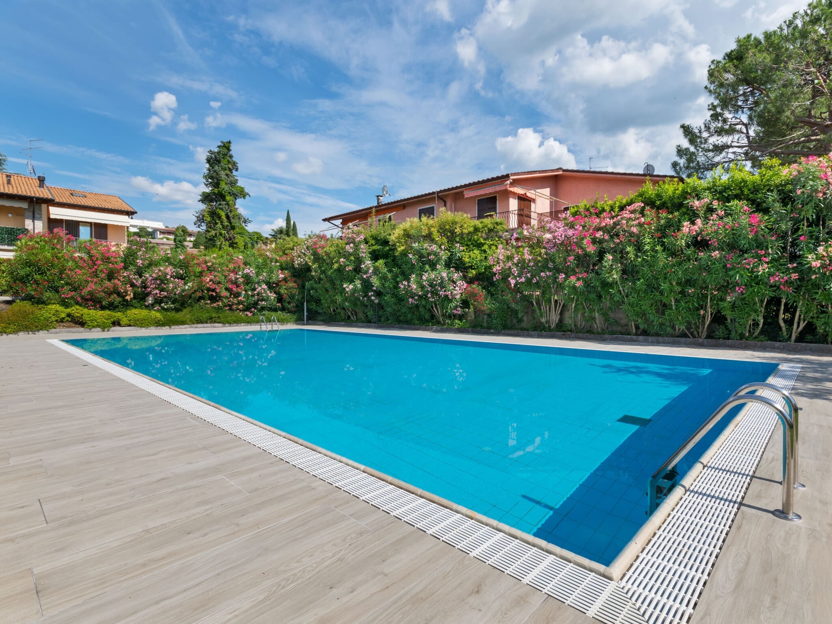 A swimming pool with a wooden deck, surrounded by lush greenery and pink flowers.