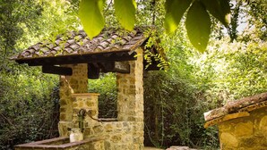 Una estructura rústica de piedra con techo de tejas, un caño y un banco de piedra.