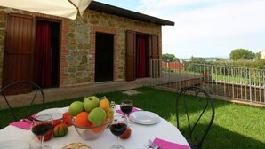 Um edifício de pedra com uma varanda, uma mesa posta para refeição com frutas e vinho, e vista para o campo.