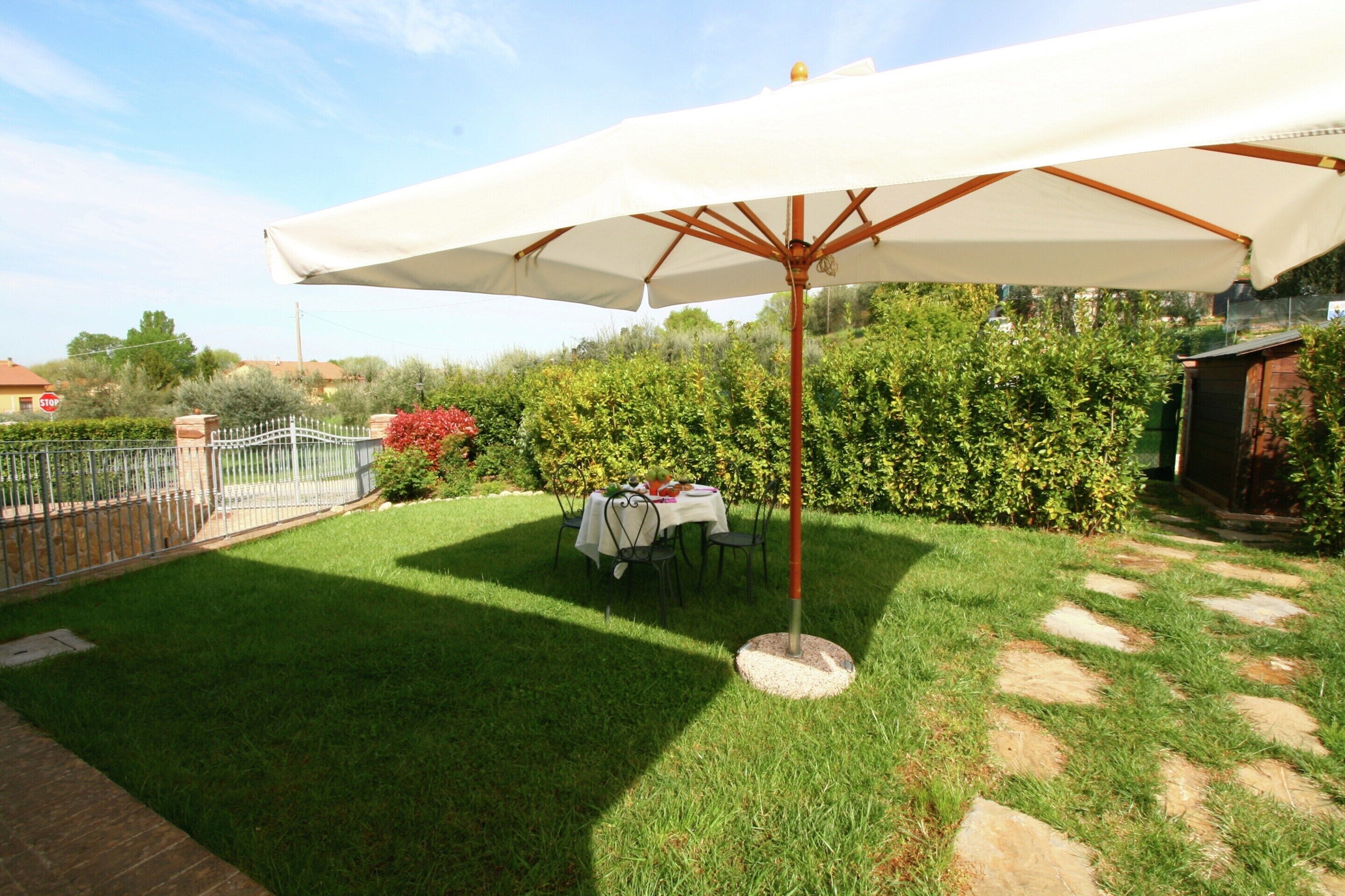 A patio area with a table and chairs under a large umbrella, surrounded by a well-maintained lawn and a hedge.