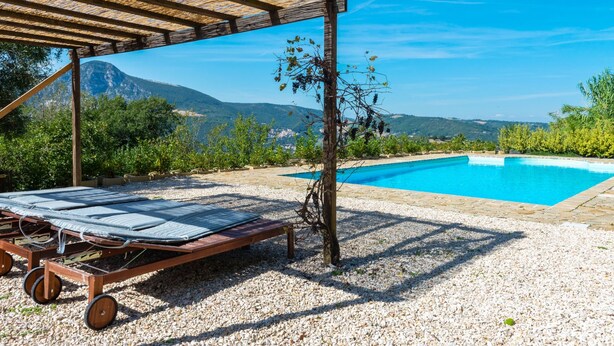 A pool area with a pergola and lounge chairs, surrounded by gravel and greenery, with a mountain view in the background. - Belvilla by OYO Villa Carina (Serra San Quirico)