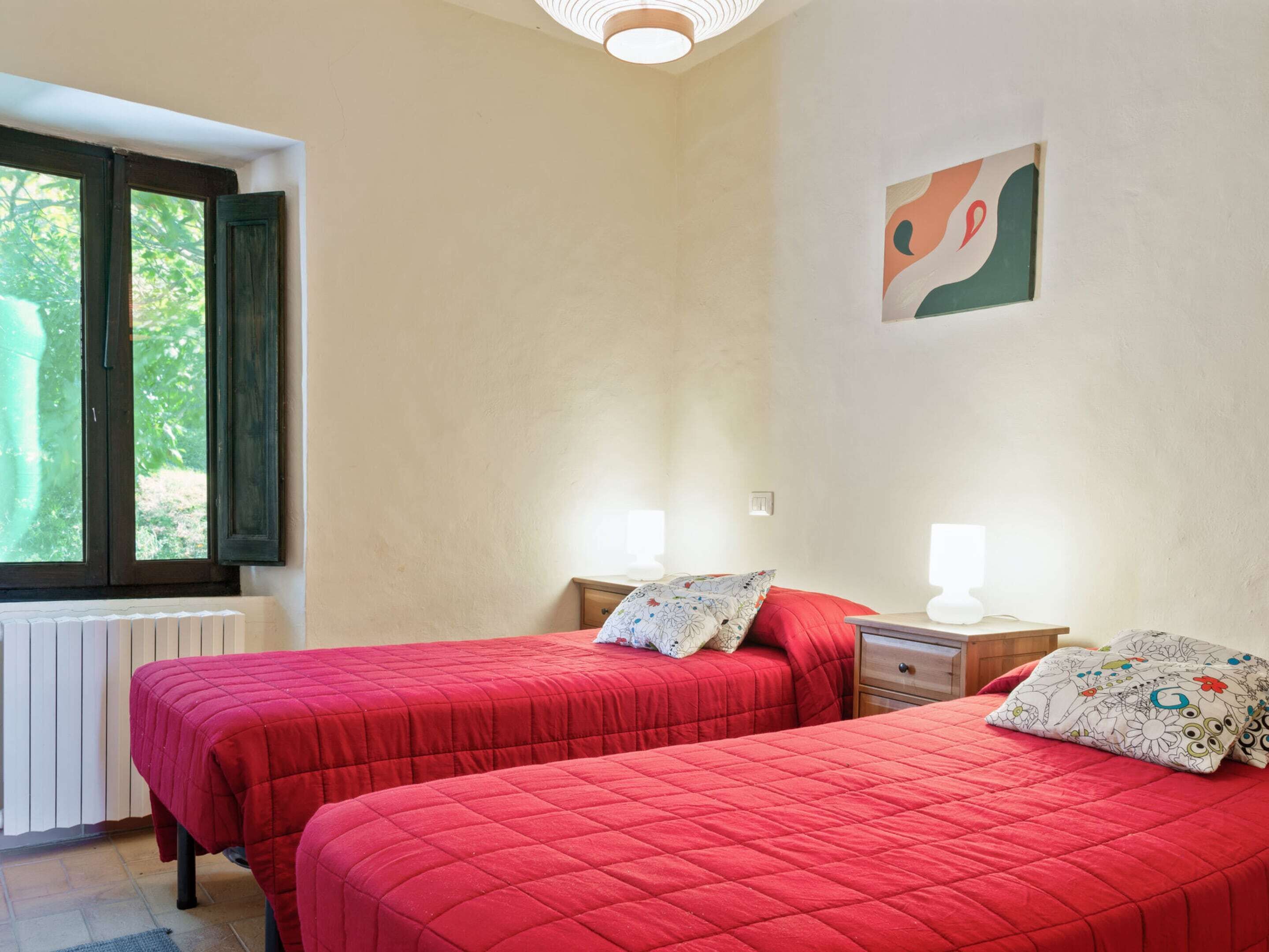 A room with two beds, each with a red quilted bedspread and matching pillows, a small bedside table with a lamp, and a window with a view of greenery.