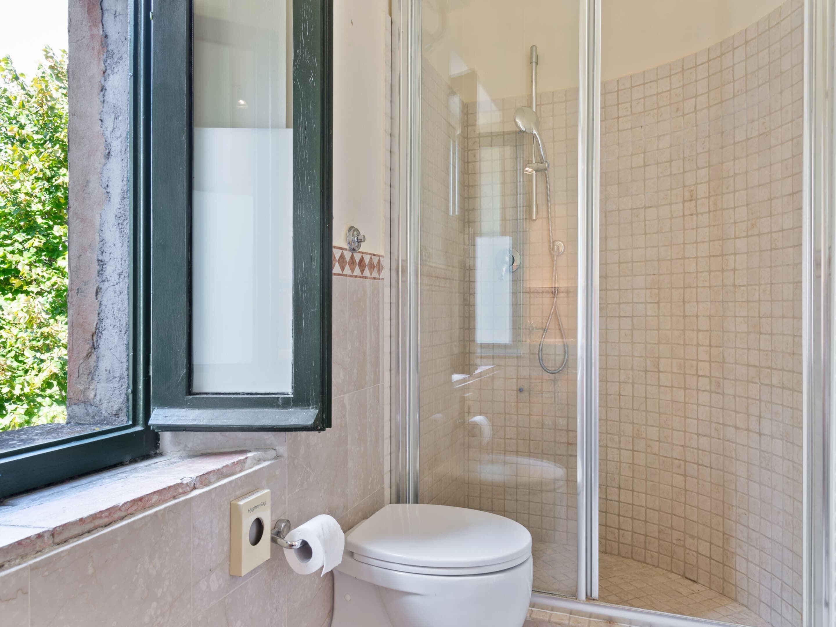 A bathroom with a toilet, a glass-enclosed shower, and a window with a view of greenery.