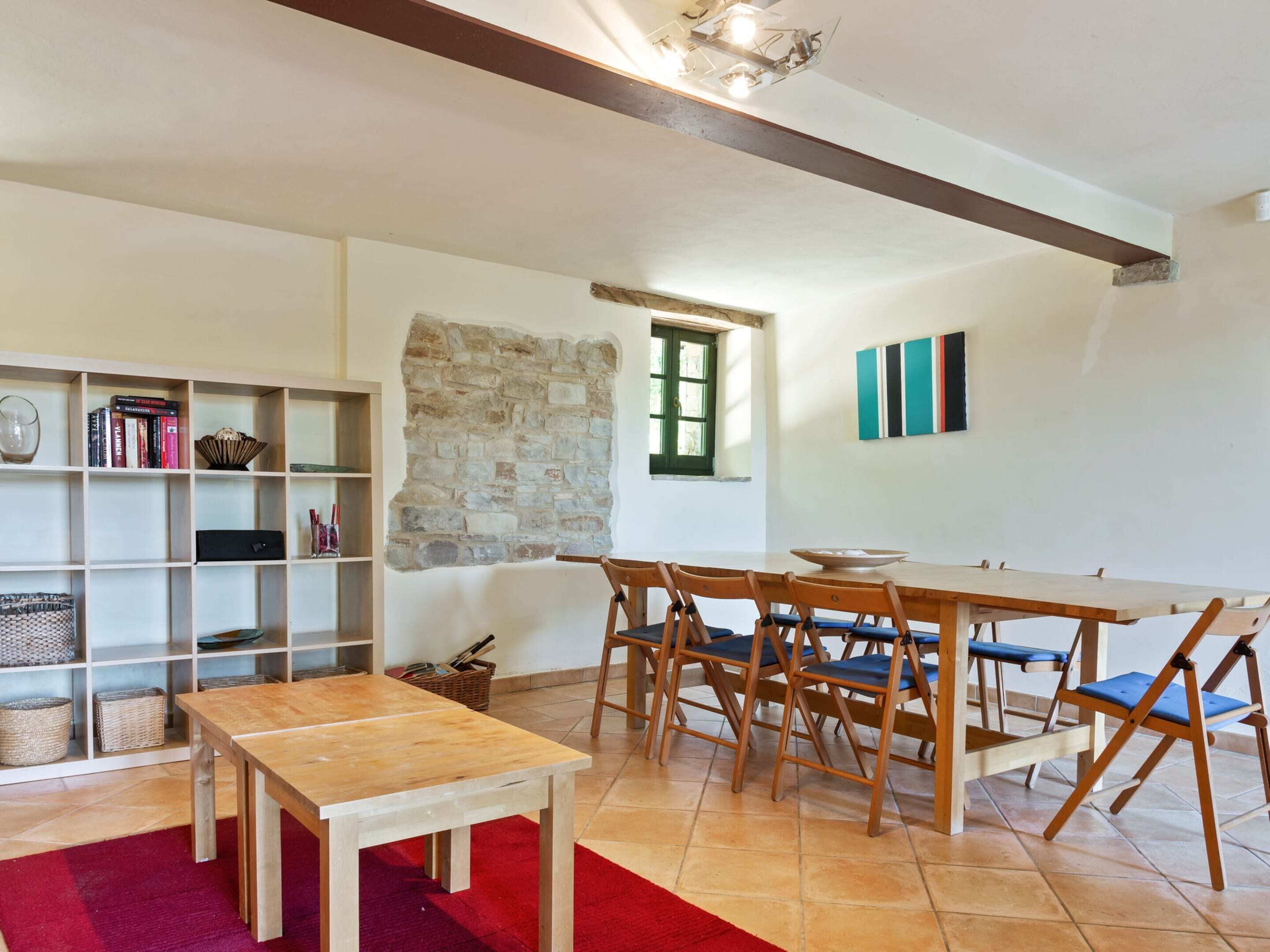 A room with a wooden table and chairs, a bookshelf, a stone wall, and a window.