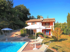 A two-story house with a swimming pool, lounge chairs, and a white umbrella.