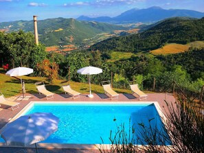 A swimming pool with lounge chairs and umbrellas, surrounded by lush greenery and a view of distant mountains.