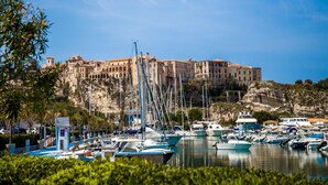 A marina with boats docked, a building with multiple stories and arches, and a rocky cliff in the background. - Belvilla by OYO Villa Conte (Ricadi)