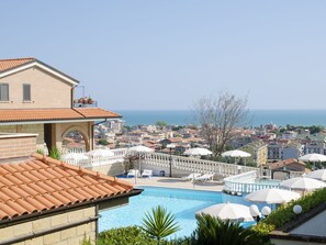 Un espace piscine avec des chaises longues et des parasols, une vue sur la mer et un paysage urbain en arrière-plan.