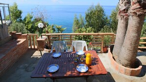 Ein Holztisch mit Tellern, Gläsern und einer gelben Karaffe auf einer Terrasse mit Blick auf das Meer.