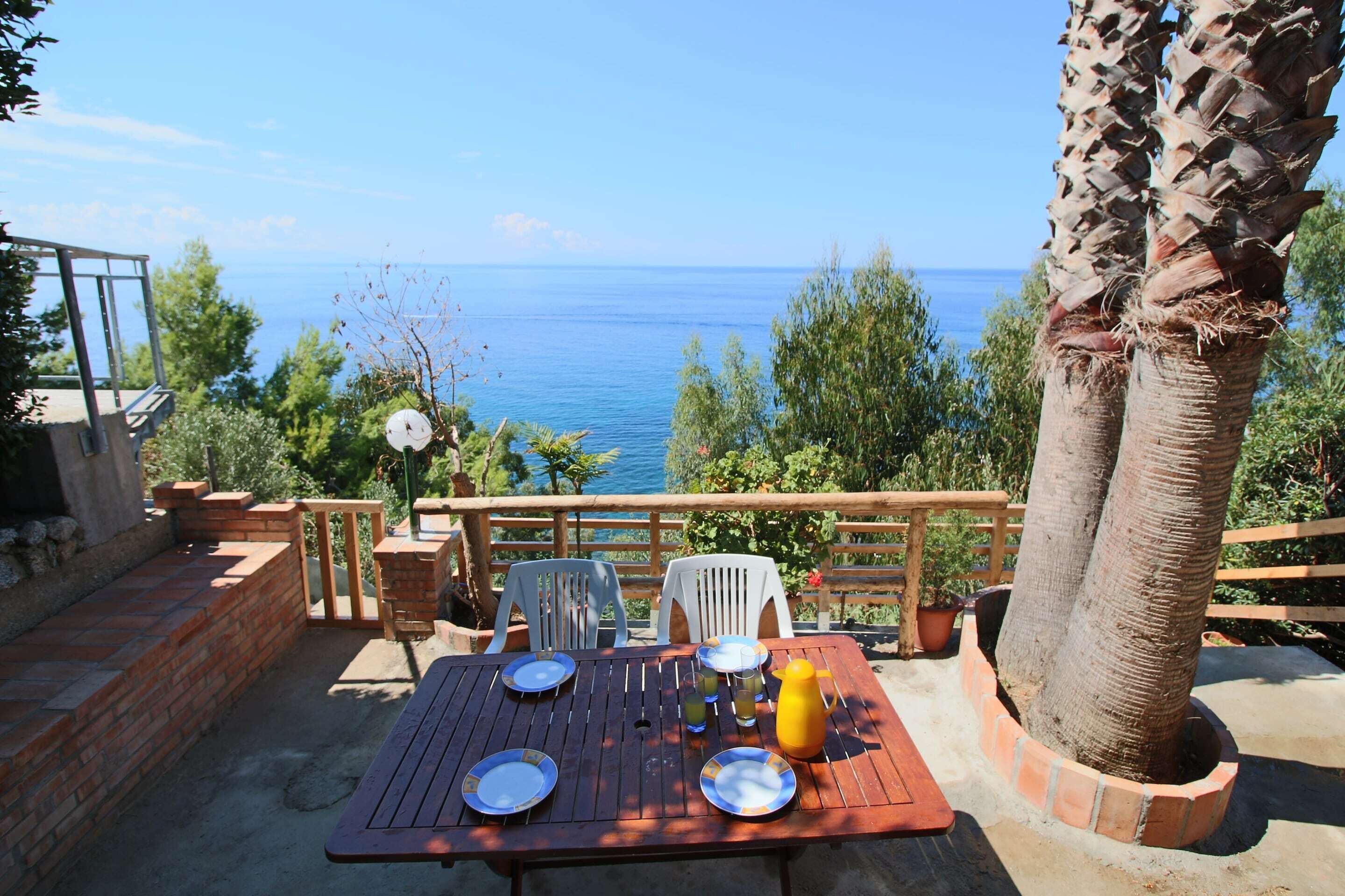A wooden table set outdoors with plates, glasses, and a yellow pitcher, overlooking a clear blue sea.