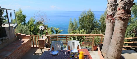 A wooden table set outdoors with plates, glasses, and a yellow pitcher, overlooking a clear blue sea.