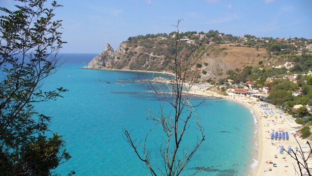 Blick auf die Küste mit Strand, klarem blauen Meer, Felsen und einem Hang mit Gebäuden.