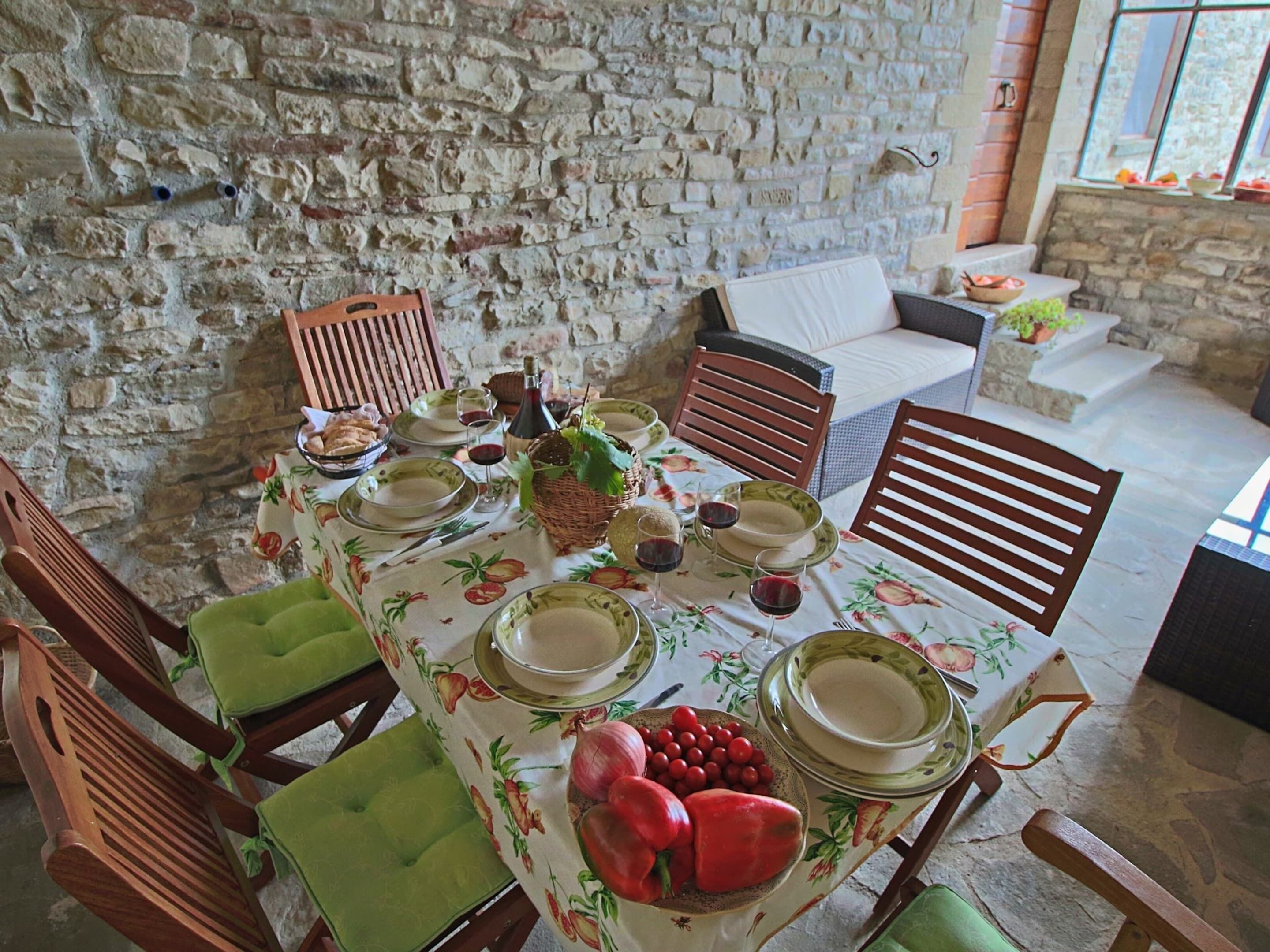 Una mesa de comedor dispuesta con platos, vasos y un centro de mesa con fruta en una habitación de paredes de piedra rústica con sillas de madera y un sofá.