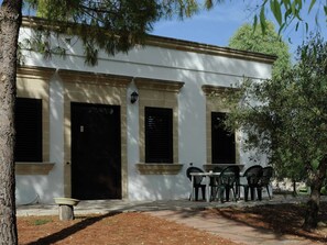 A building with three black doors and windows, a small outdoor seating area with plastic chairs, and a tree in the foreground.