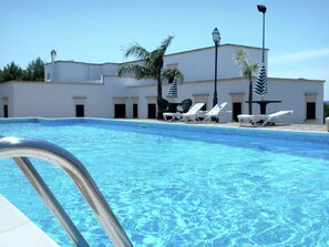 A swimming pool with clear blue water, surrounded by white lounge chairs and umbrellas. There are palm trees and a building with a white facade in the background.