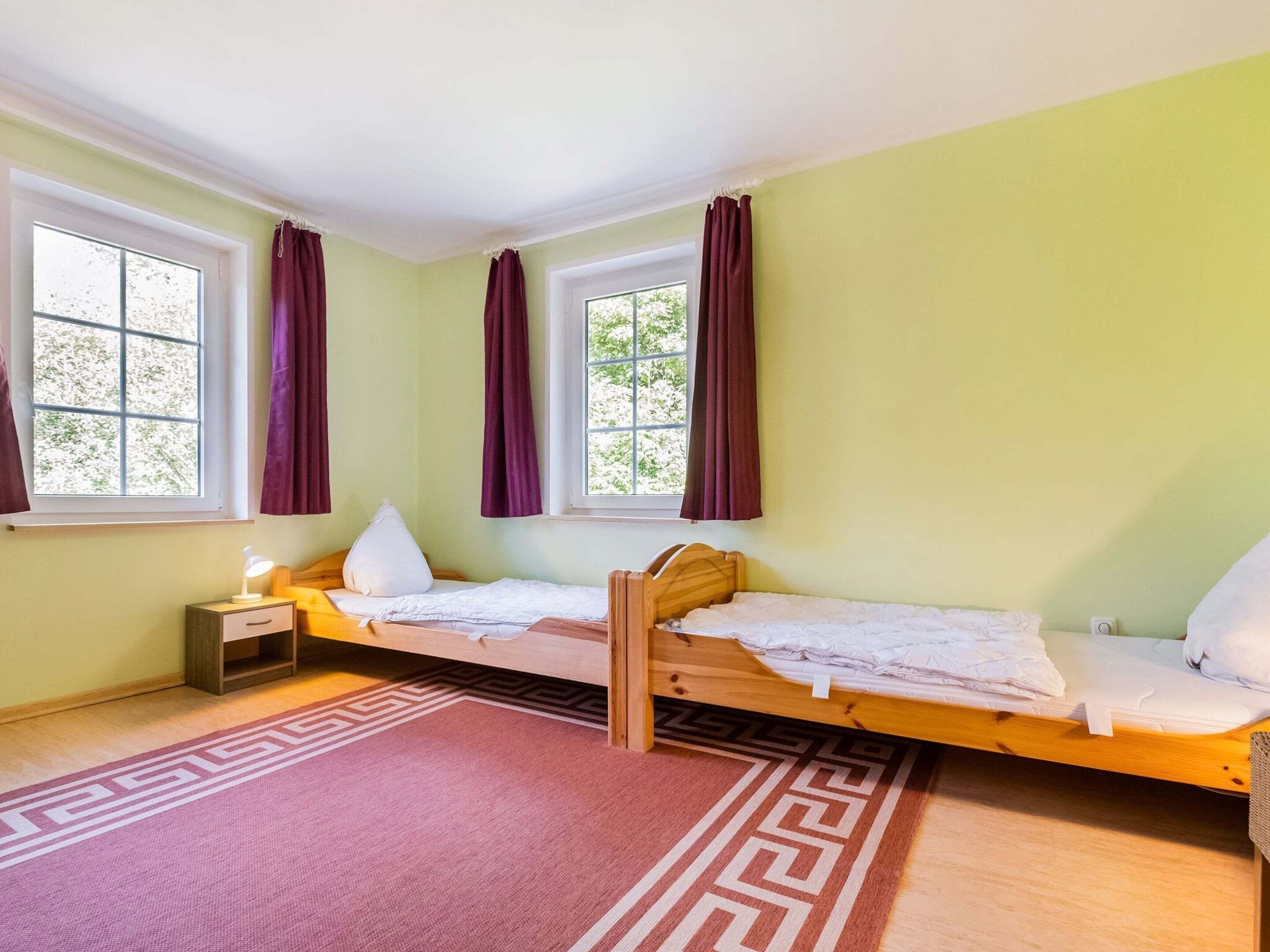 A small, single-bed room with a window, a bedside table with a lamp, and a patterned rug.