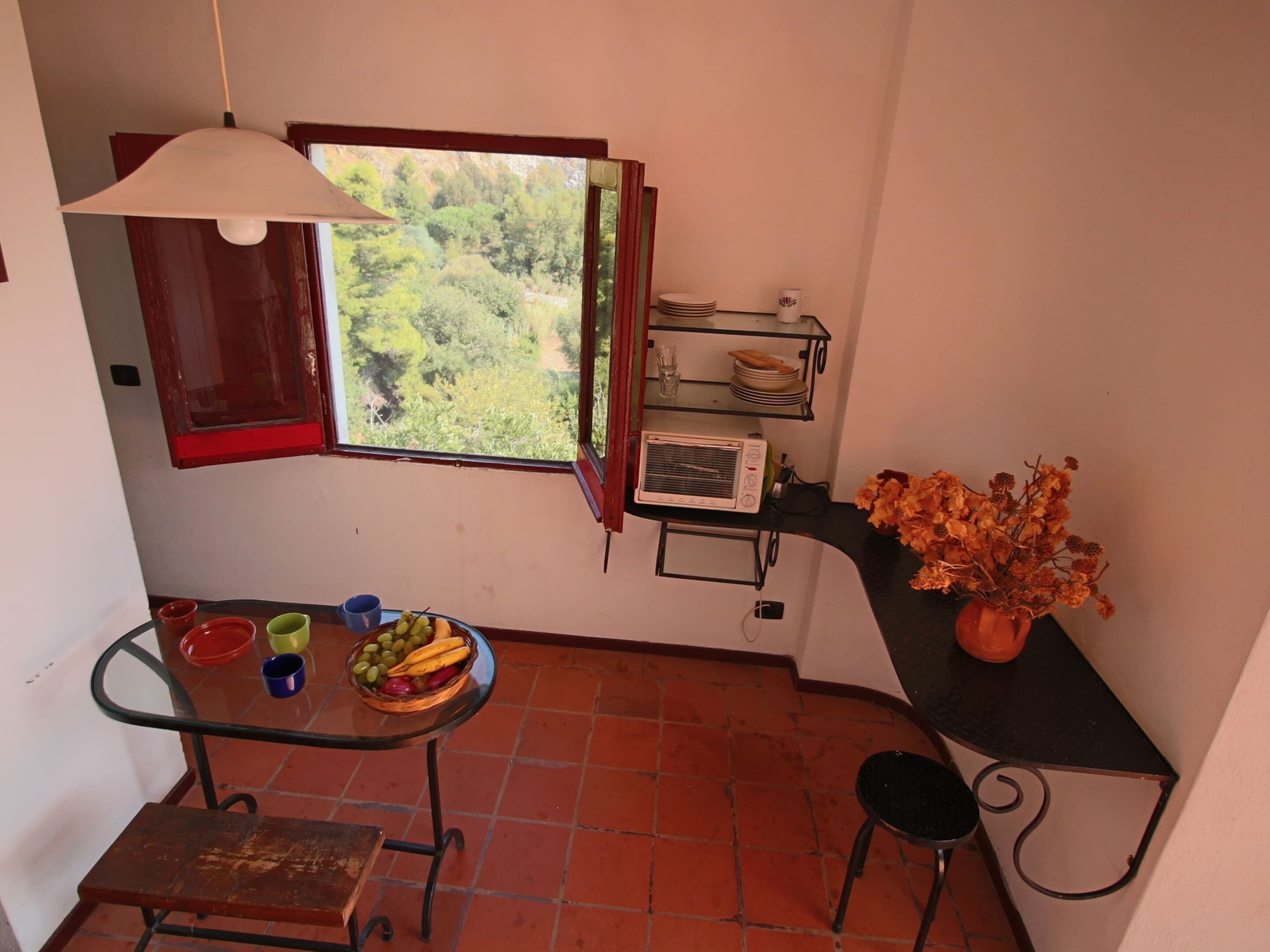 A compact kitchen with a window, a table with fruit, a small stool, and a decorative vase.