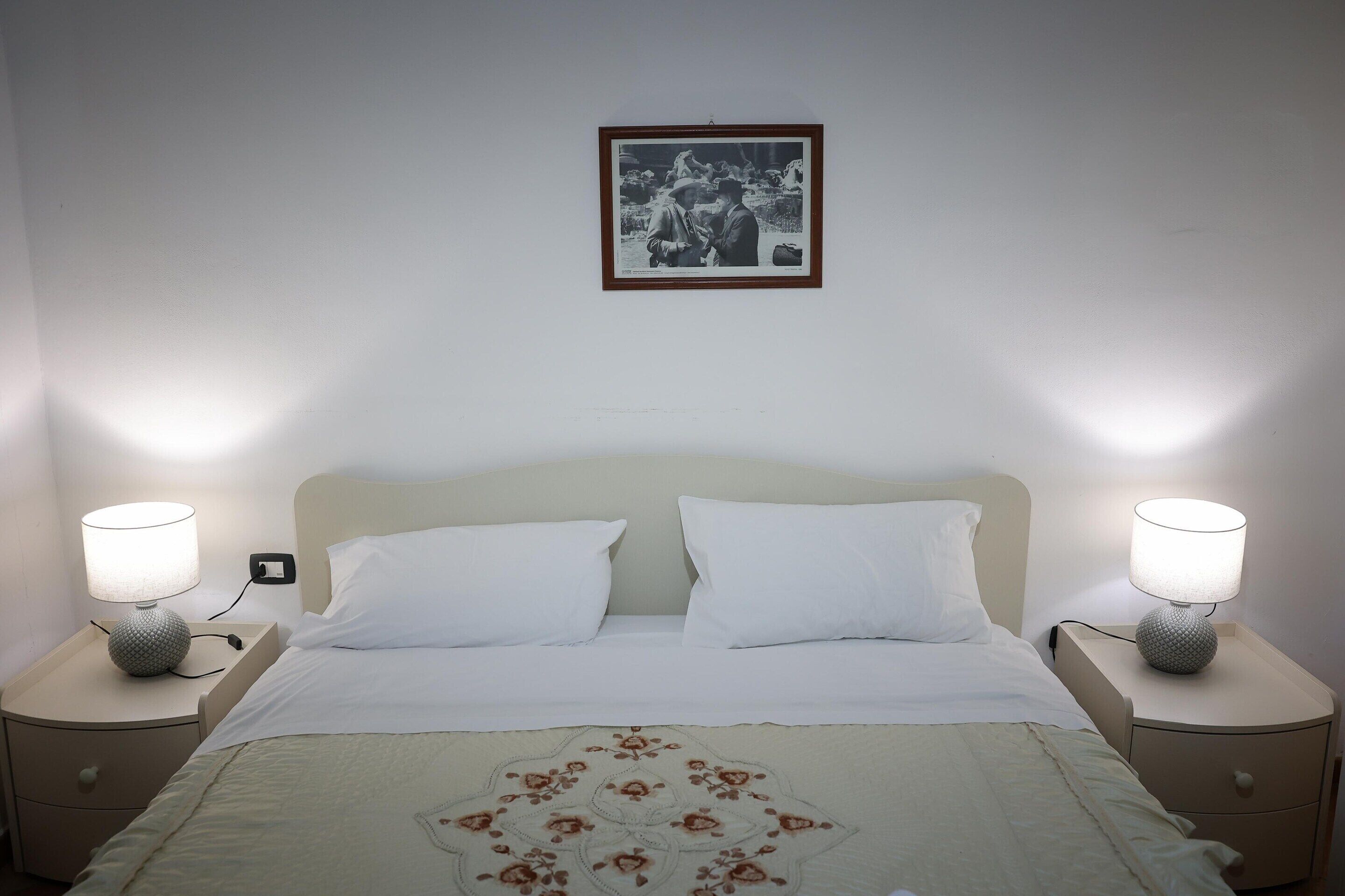 Ein ordentlich bezogenes Bett mit weißen Leinen und einer floralen Tagesdecke, neben dem sich zwei Nachttische mit Nachttischlampen befinden. Ein gerahmtes Bild hängt über dem Bett.
