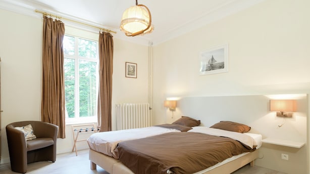 A bedroom with a large bed, two bedside lamps, a chair, a window with curtains, and a radiator. - Captivating Holiday Home in Malmedy With Sauna (Malmedy)