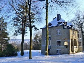 Ein steinernes Gebäude mit schneebedecktem Dach und zahlreichen Fenstern, umgeben von Bäumen und einer verschneiten Landschaft.
