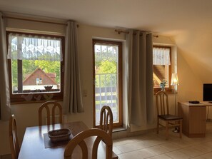 Ein Zimmer mit Esstisch, Stühlen, einer Lampe, einem Fernseher und Fenstern mit Vorhängen.