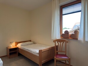 Ein Einzelbett mit Holzrahmen, ein Nachttisch mit Lampe, ein Sessel mit gepolstertem Sitz und ein Fenster mit Jalousien.