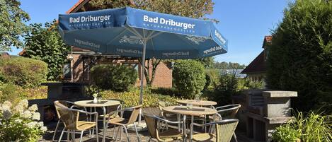 Ein Außenbereich mit Tischen und Stühlen unter einem blauen Sonnenschirm mit dem Schriftzug 'Bad Driburger'.