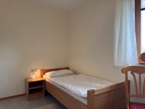 A small, single-bed room with a wooden headboard, a bedside table with a lamp, and a window with a white curtain. - Holiday Apartment in the Teutoburg Forest (Brakel)