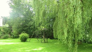 Um jardim com balanço, casinha de madeira e um salgueiro-chorão.