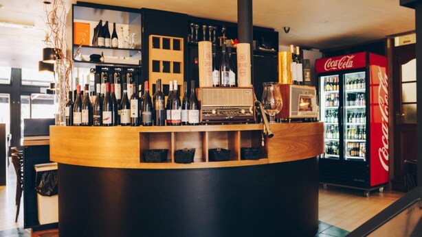 Un coin bar avec un comptoir en bois, des étagères remplies de bouteilles, une radio ancienne et un réfrigérateur Coca-Cola.