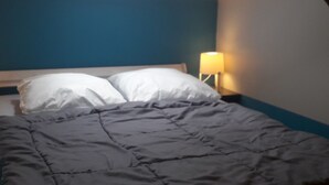Ein Bett mit dunkelgrauer Decke und weißen Kissen, ein Nachttisch mit Lampe und eine blaue Wand.
