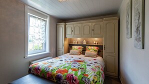 Quarto com colcha florida, janela com persianas, um banco e uma prateleira decorativa com objetos.