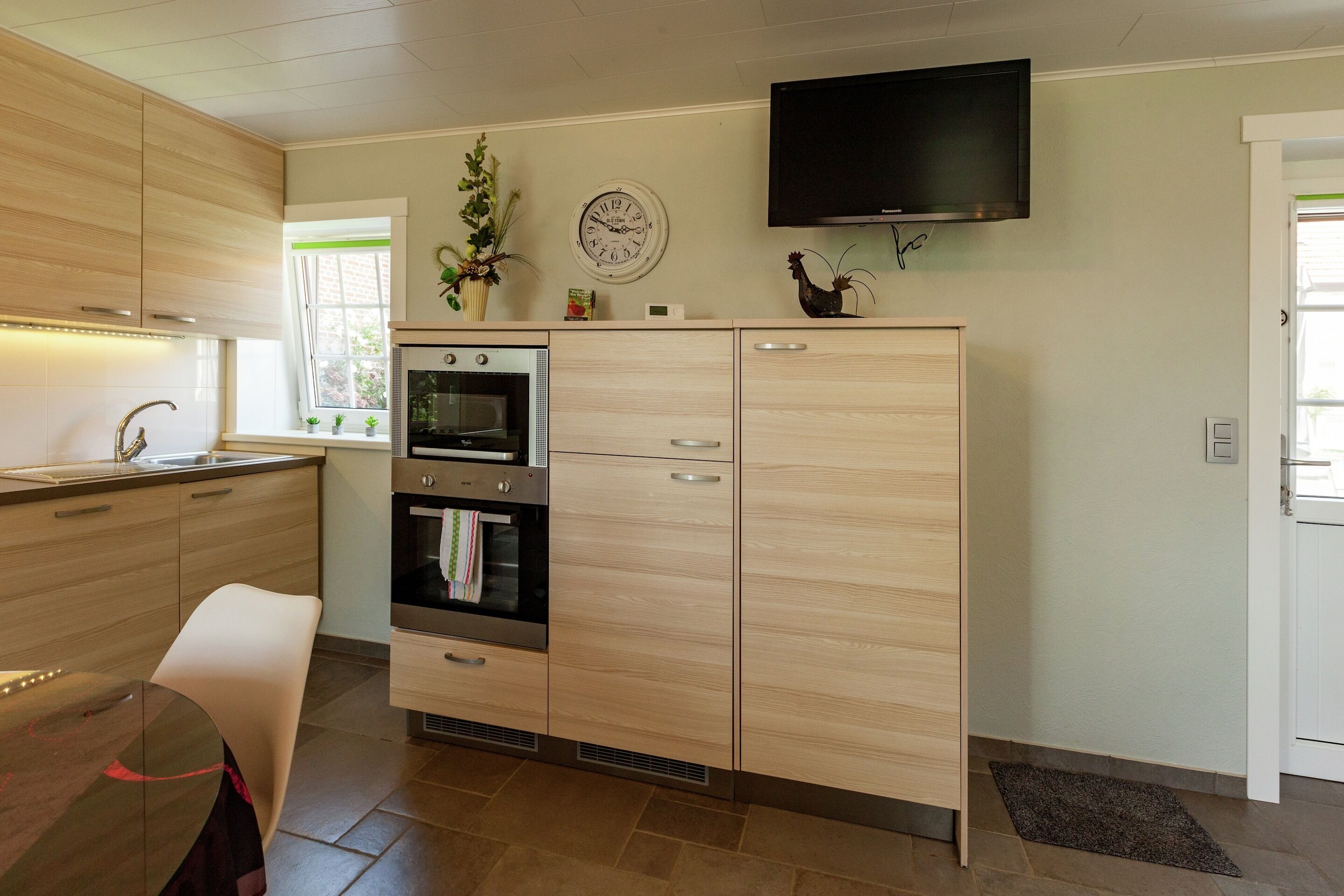 A modern kitchen with wooden cabinets, a built-in oven, a microwave, and a flat-screen TV mounted on the wall.