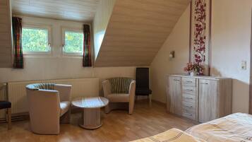 A room with a sloped ceiling, a bed, a sofa, a chair, a small table, a cabinet, and two windows with curtains.