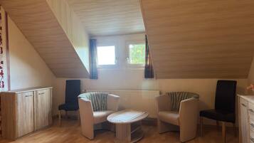 Quarto com teto inclinado, mesa redonda, duas cadeiras, radiador e janela com cortinas.