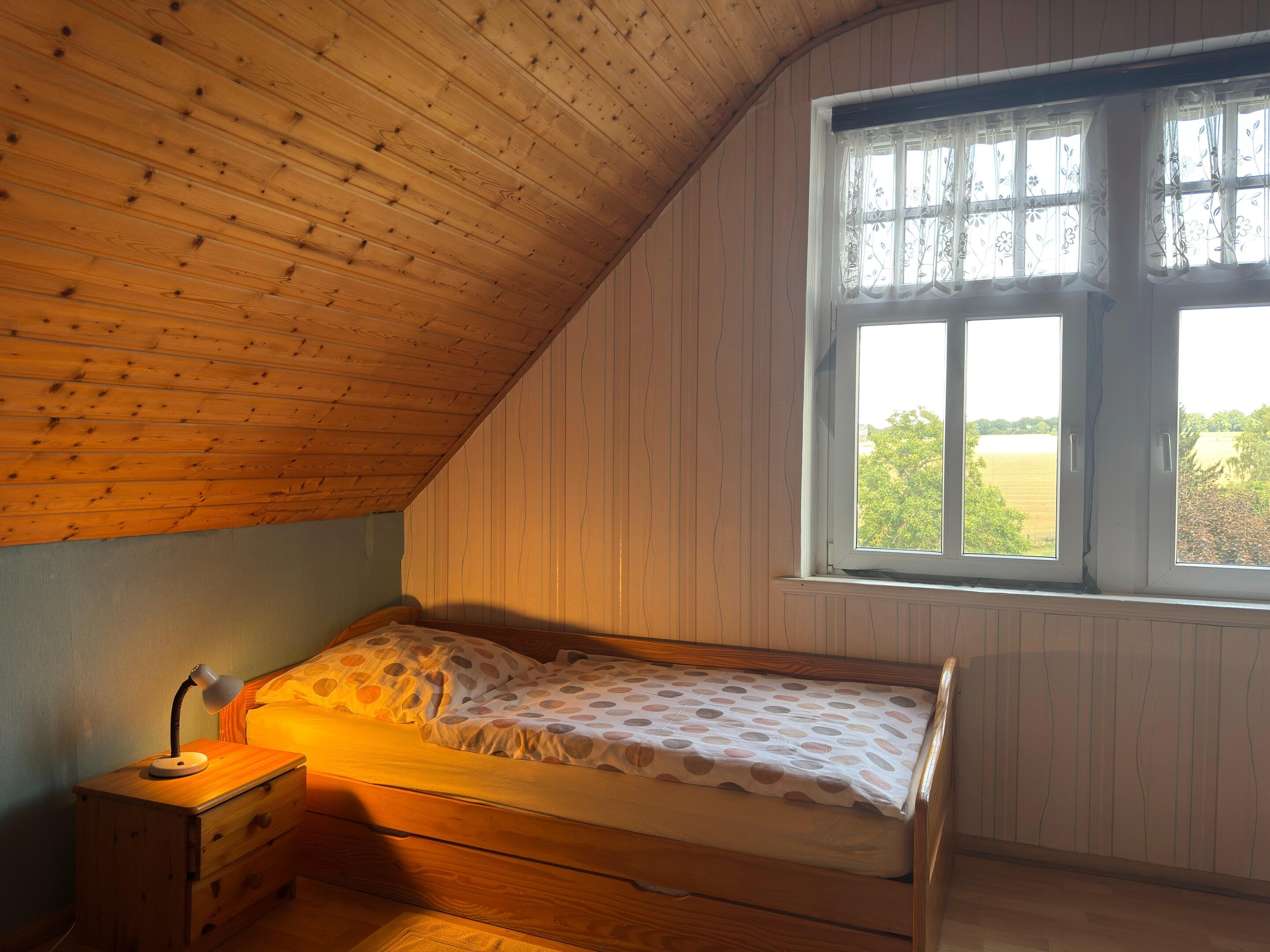 Un dormitorio con techo inclinado, cama de madera, mesita de noche con lámpara y ventana con vistas a follaje.