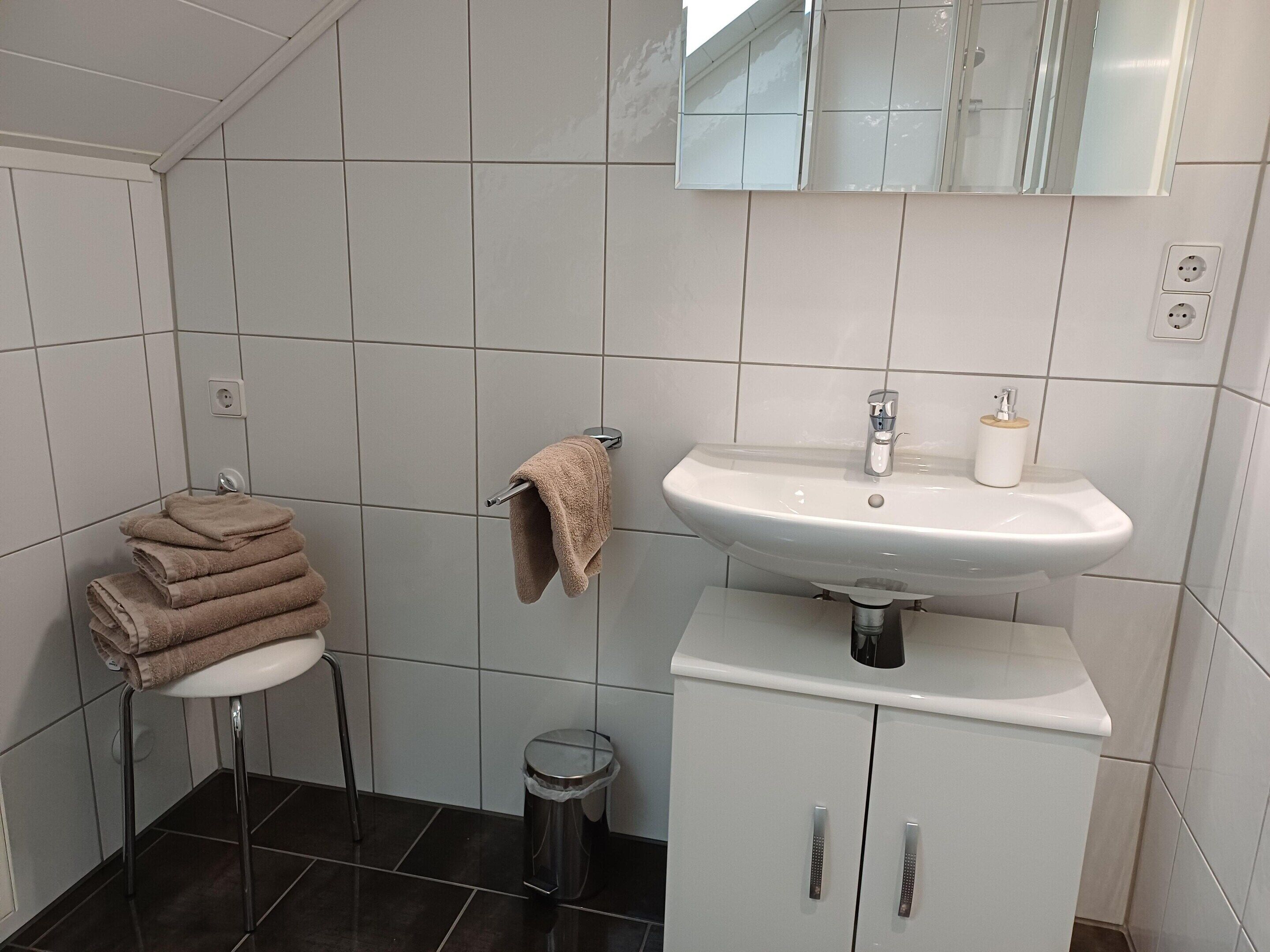 A bathroom with a white sink, mirror, and tiled walls. There is a towel rack with folded towels and a trash can.