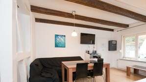 Um quarto com uma mesa de jantar de madeira e cadeiras, um sofá preto, uma televisão fixada na parede e uma janela com vista para o verde.