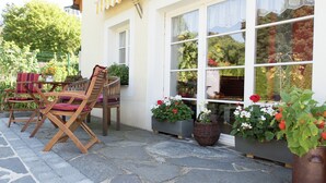 A patio with wooden chairs and a table, large windows with lace curtains, and potted plants. - Comfortable Holiday Home in Saxony With Terrace (Oederan)