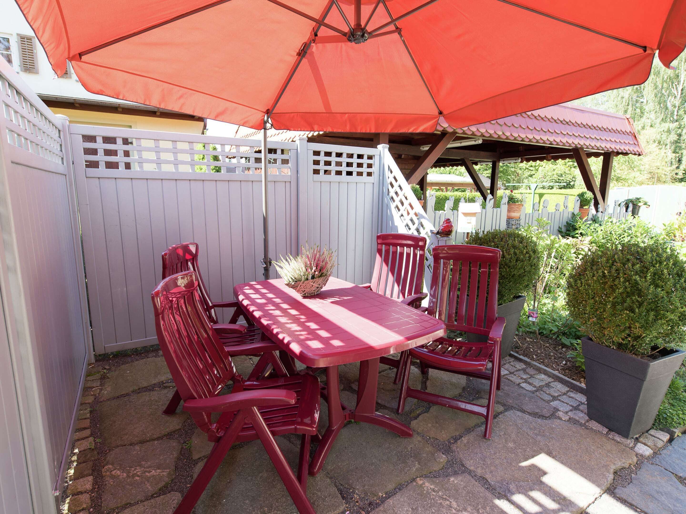 A patio with a red table and chairs, a red umbrella, and a white fence.