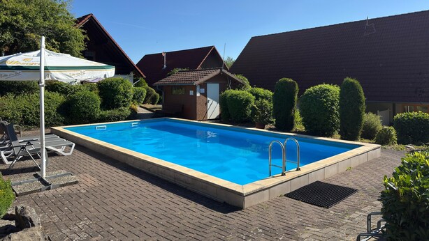 Piscina retangular com deck revestido de azulejos, cercada por vegetação e um pequeno edifício com telhado inclinado.