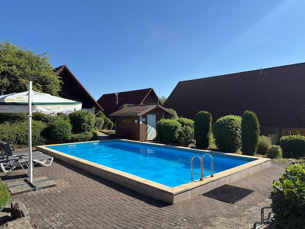 A rectangular swimming pool with a tiled deck, surrounded by greenery and a small building with a sloped roof. - Holiday Home in Germany With Garden Terrace (Brakel)