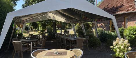 Un espacio de comedor al aire libre con mesas y sillas bajo una carpa, rodeado de follaje y un edificio de ladrillo.