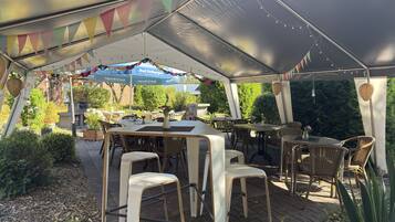 Eine Terrasse mit Tischen und Stühlen, ein überdachter Bereich mit Zelt und ein Garten im Hintergrund.