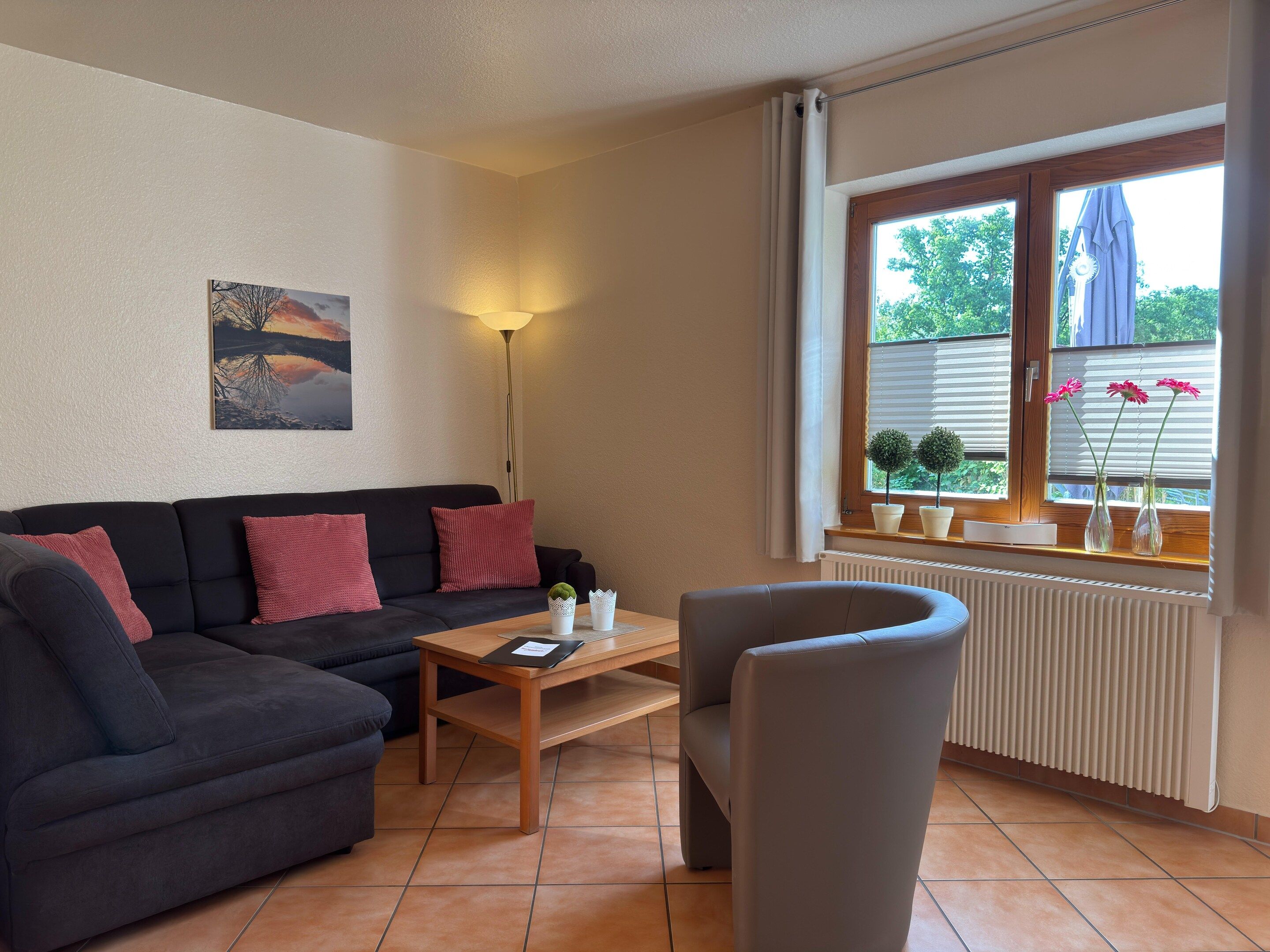 Una sala de estar con un sofá, una mesa de centro y un sillón. Hay una ventana con persianas y un radiador debajo.