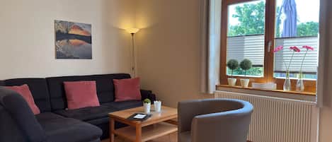 Una sala de estar con un sofá, una mesa de centro y un sillón. Hay una ventana con persianas y un radiador debajo.