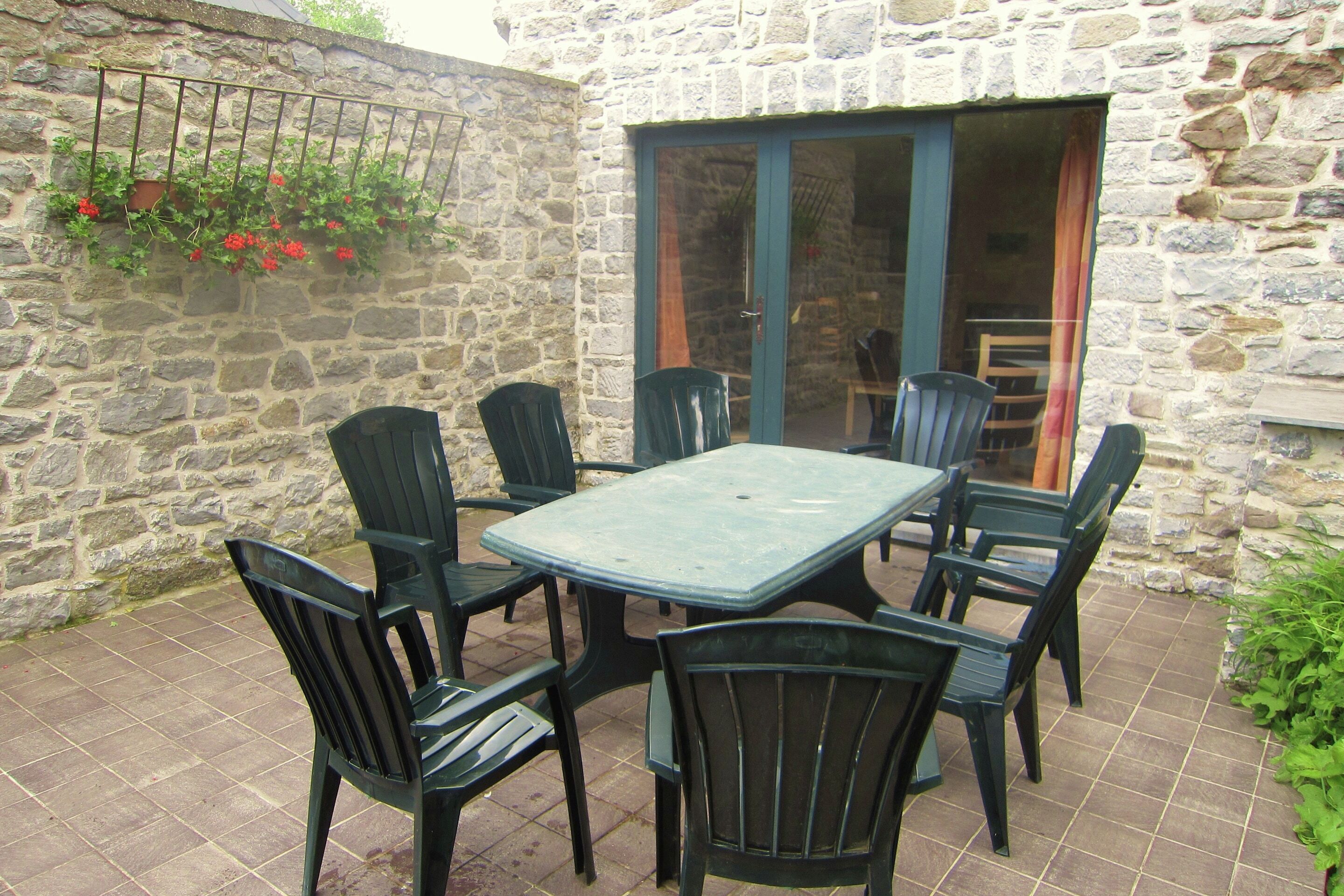 Eine Terrasse mit Steinwänden, einem Tisch, Stühlen, einer Schiebetür und einem blumengeschmückten Korb.